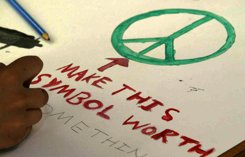 The CND symbol (or peace symbol) being drawn by child with the words 'make this symbol worth something' underneath