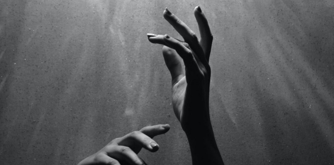 A black and white image of two hands reach up out of the dark to the light