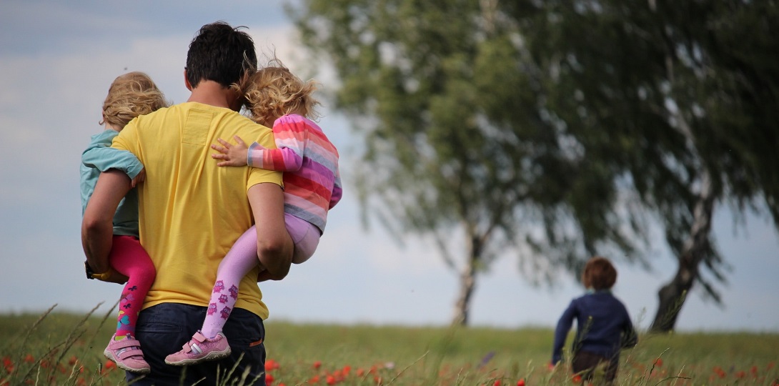 A man carrying two young children through a grassy field