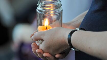 A candle in a jar clasped in a persons hand. Further candles held by people can be seen blurred in the background