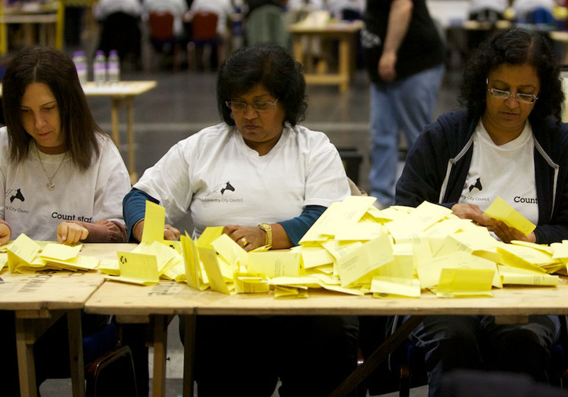Three women seated at table counting hundreds of yellow ballot papers