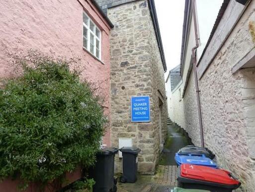 Stone house partially viewable from narrow alleyway, blue sign reads Quaker Meeting House. 