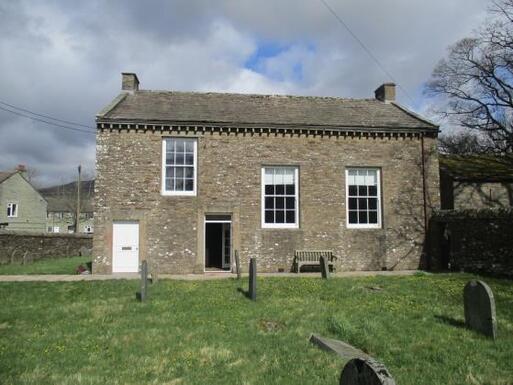 Short, squat stone building with large windows, situated within a burial ground. 