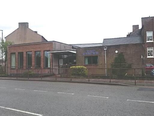 Flat roofed brick building with five long windows, sloping covered entryway, and 'Quaker Meeting House' lettering on the right side fascia. 