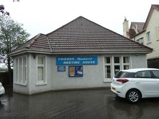 White bungalow within a car park with noticeboards and blue 'Friends (Quakers) Meeting House' sign affixed to the building. 