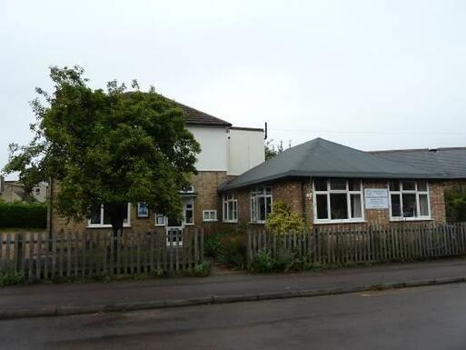 Two storey brick house with large front extension and fenced garden, white sign attached to front fascia.