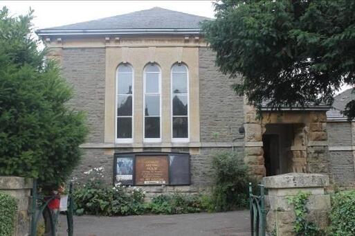 Tall stone building with three long windows to front, a large noticeboard sits underneath these and to the left of the large raw-stone entrance. 