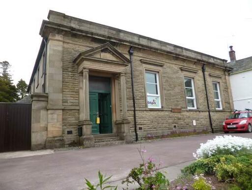 Old stone building with columned portico, large long windows, and small car park to front. 