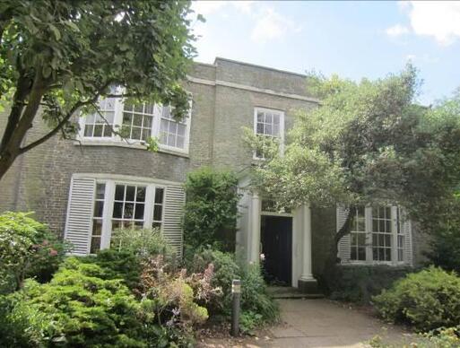 Large brick building with four large bay windows surrounding main front door, all set within lush gardens. 