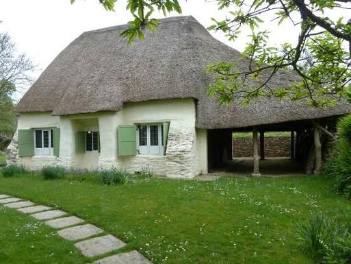 Small cottage with large thatched roof and three windows flanked by light-green shutters. 