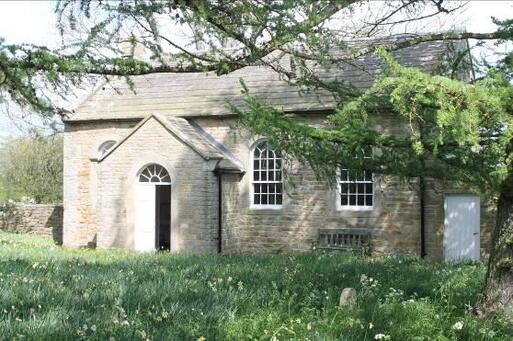 One storey stone building with large arched windows and entryway porch, set within large meadow gardens.