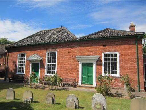 Small redbrick building with long white windows and dual large green doors, set within a small Quaker burial ground. 