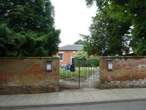 Short brick building set in large gardens is viewed through metal gate set in tall brick boundary wall.