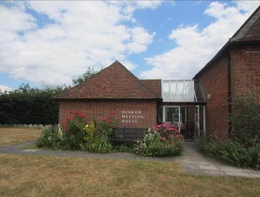 Square brick meeting house is joined to a house by a glass extension, 'Quaker Meeting House' lettering on its front wall, all set within large garden grounds.