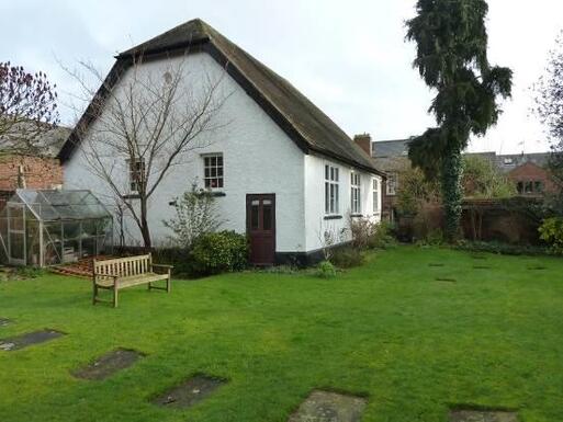 White cottage with tall roof set within large garden grounds.