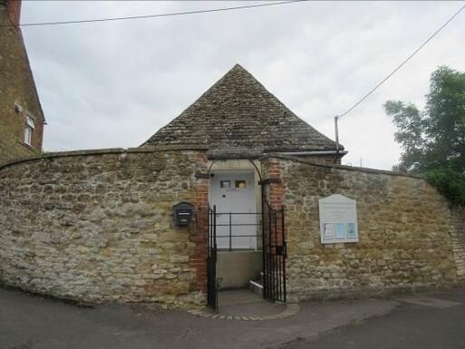 Small old stone building is nearly completely obscured by a high stone boundary wall, a white front door is visible through the entrance gate.