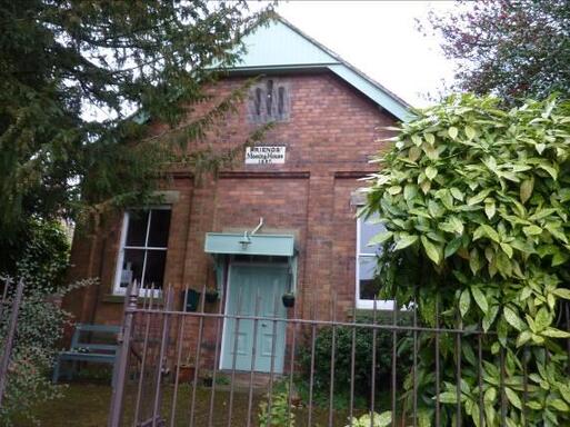 Short brick building with light green doors and two white windows, behind lush garden hedges. 