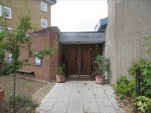 Large concrete building with brick side-entrance topped with grey sign reading 'Quaker Meeting House'.