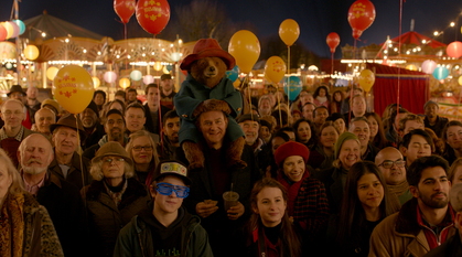 A bear in clothes sits on middle-aged man's shoulders in a crowd of people with balloons, all looking at something.