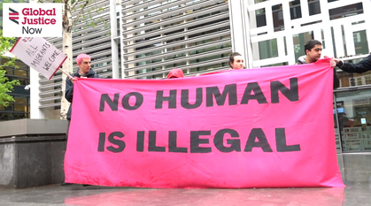 5 people holding up a bright pink banner outside on a grey day with 'No Human Is Illegal' on it. 