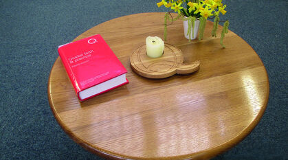 A picture of a wooden table with a handmade 'Q' candle holder, flowers, and Quaker Faith and Practice