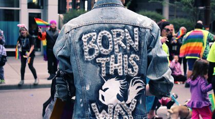 Person wearing a denim jacket with 'born this way' written on the back with a rampant unicorn, standing by the side of family-friendly pride event with lots of rainbow flags around.