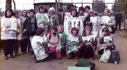 Quaker women outside the fence at RAF Greenham Common wearing and carrying posters and banners about supporting peace and against dropping bombs.