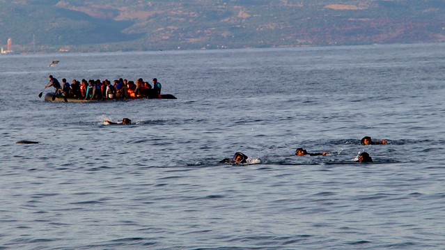 People crowded into a small boat on the sea. Some people swimming around it