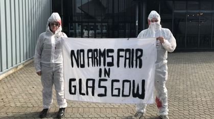 two people in 'blood' spattered hazmat suits holding a banner saying 'no arms fair in Glasgow'