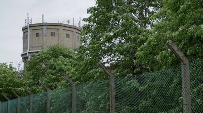 a chain link fence in front of some leafy trees. A watchtower looms in the background.