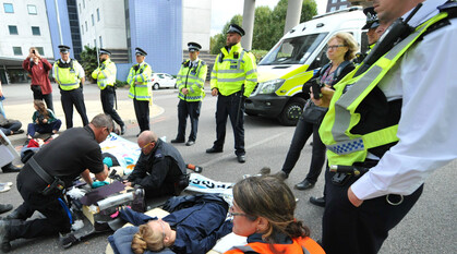 protestors laying across the road in a lock on protest at an anti-arms fair demo in London. They are surrounded by police and two police officers are cutting through the suitcase being used for the lock on
