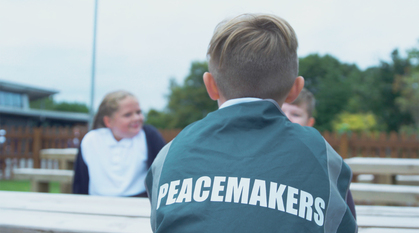 three children sat around a bench outside. One is wearing a tabard that says peacemaker