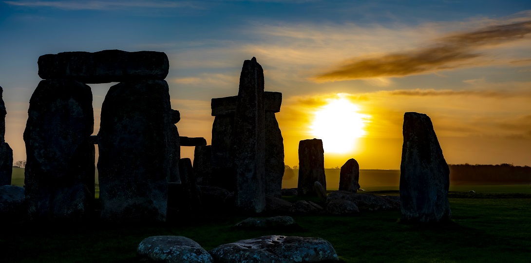 Silhouette of an ancient tone circle against a dark blue sky with the sun setting in the background