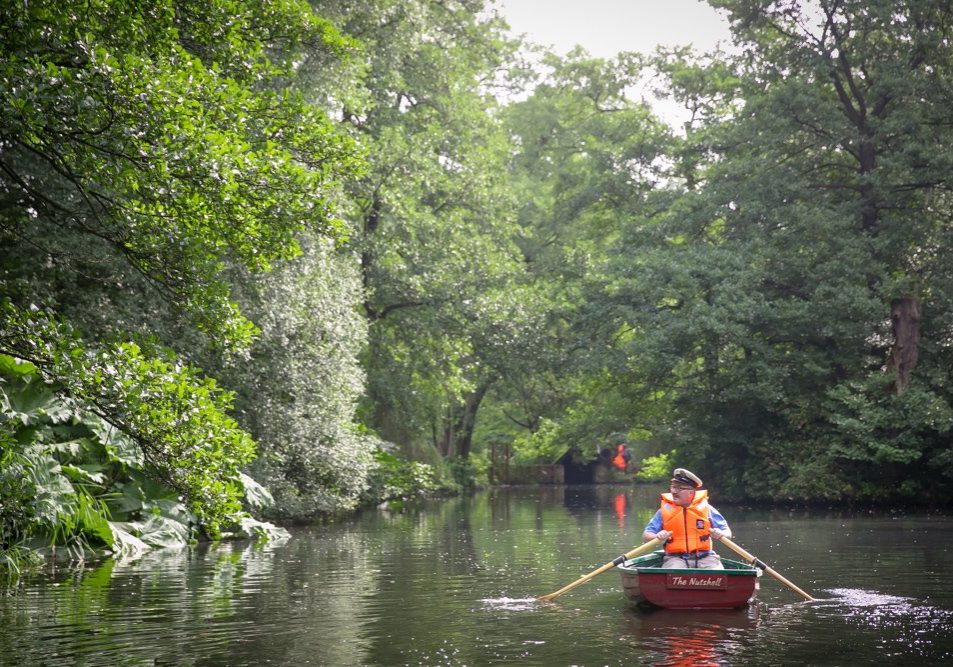 A photo of a man in a sailor's cap and orange lifejacket rowing a small boat on a pond surrounded by trees