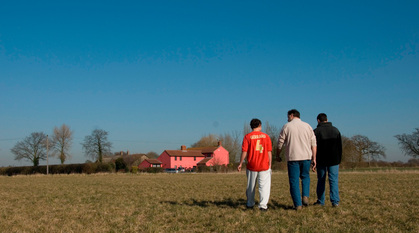 Three people walking across a field towards a pink cottage glowing in the sunlight underneath a blue sky