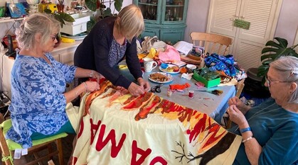 3 Friends stitching together a banner at a dining room table in someone's home