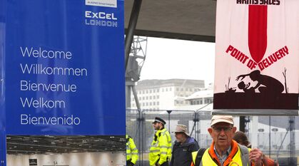 Next to a 'Welcome to the ExCel Centre' sign, a Quaker stands with a banner saying 'arms sales -> point of delivery'. The image on the banner is a simple silhouette drawing of a parent and baby with a rocket coming down on them