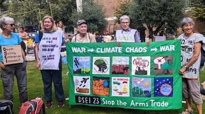 A group of Quakers wearing protest placards and tabards holding up a handmade banner with images depicting how war leads to climate chaos which leads to more war