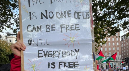 Person in a Quakers for climate justice t-shirt holding up a sign at a ceasefire rally with a quote from Maya Angelou "The truth is no one of us is free until everybody is free"