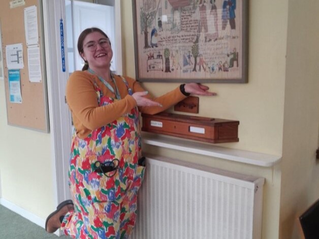 Julia is smiling and wearing colourful dungarees while standing in front of a tapestry of Chichester meeting house.