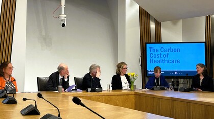 A panel of speakers in a room in the Scottish Parliament. A screen behind the speakers says 'The Carbon Cost of Healthcare'