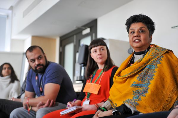 Close up of three people who are sitting in a circle in a workshop. One person is in focus wearing a bright yellow scarf with blue embroidery. She is speaking.