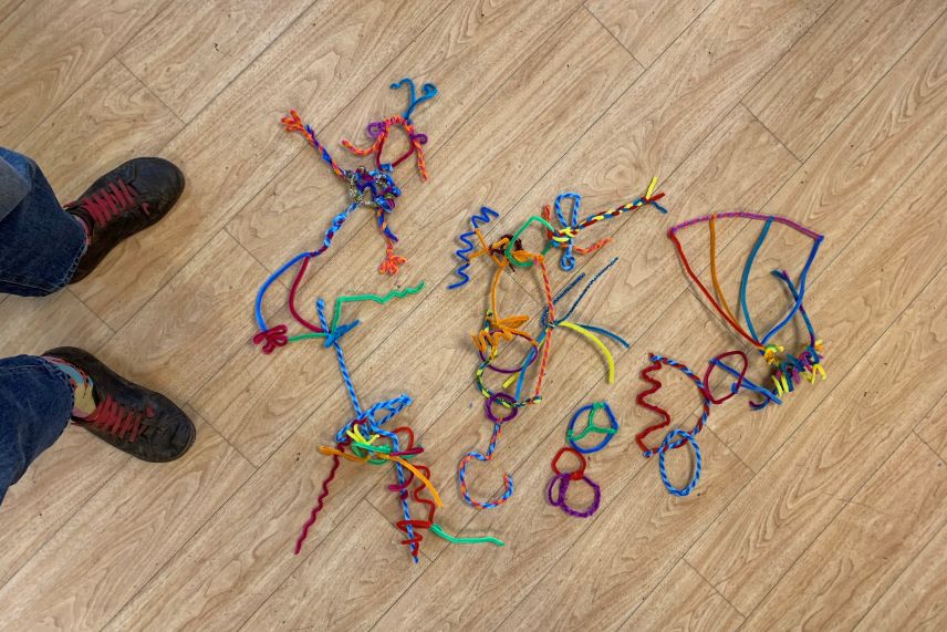 persons feet and shoes, coloured pipecleaners twisted into shapes on a wooden floor
