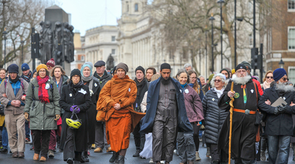 People in a variety of religious dress wearing or carrying white flowers walking silently down Whitehall. The Women of World War II memorial is visible in the background.