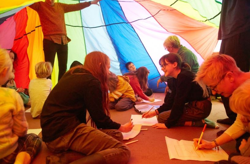 adult and children sat on the floor talking and writing under a colourful parachute