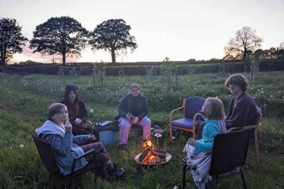 group of young people sat on chairs around a camp fire in a field with trees