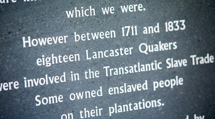Focus on grey plaque in Lancaster Meeting House. Plaque reads 'Quakers are known as slave trade abolitionists, which we were. However, between 1711 and 1833, 18 Lancaster Quakers were involved in the Transatlantic slave trade. Some owned enslaved people on their plantations. ...'