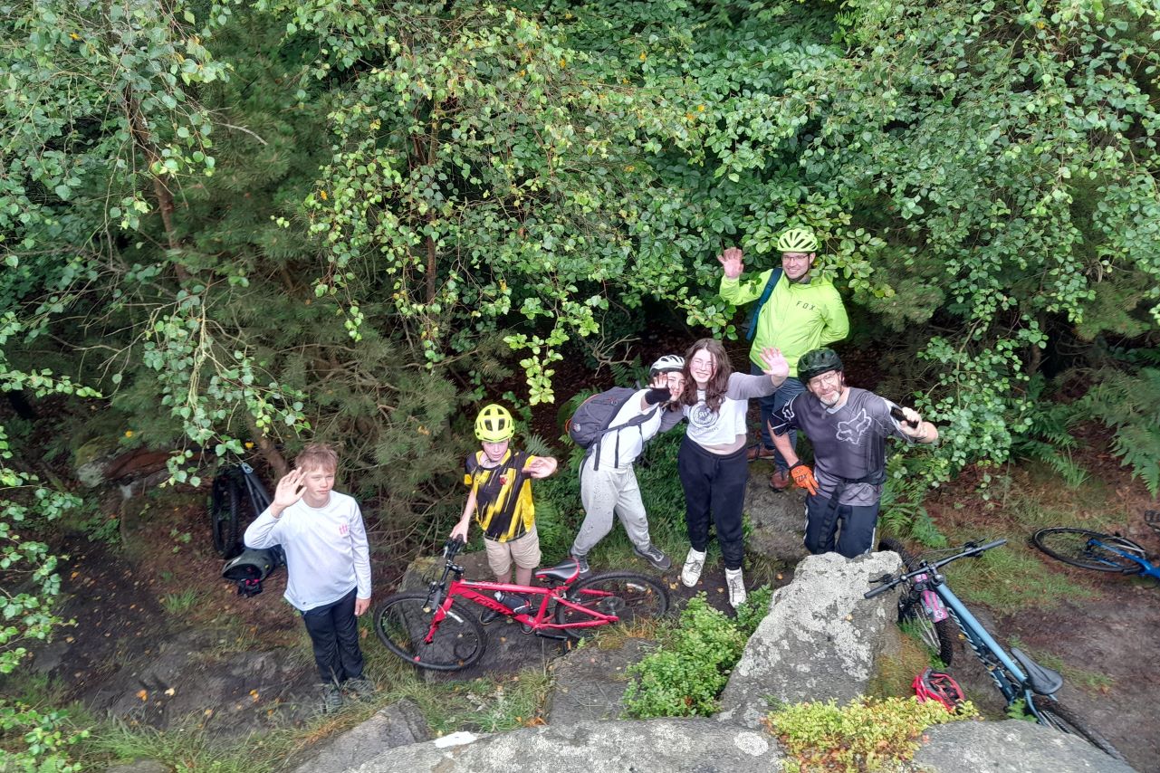 group of 6 people wave at camera standing beside bikes in woods. 