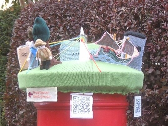 knitted postbox topper with figures and building on top of red post box