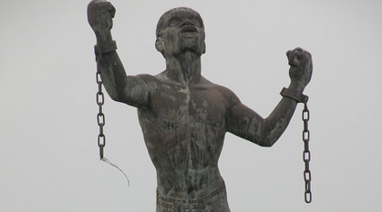 Sculpture of a Black man standing strong with face and arms raised upwards, the manacles around his wrists broken apart